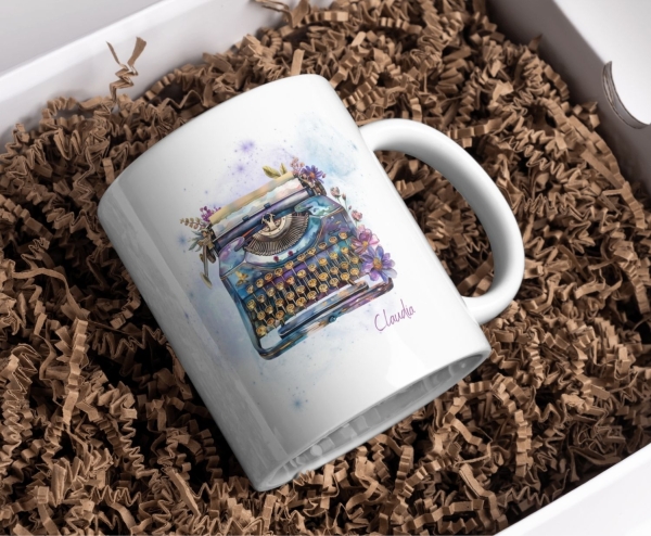 Weiße Keramiktasse mit einer kunstvollen Aquarell-Illustration einer Vintage-Schreibmaschine, verziert mit floralen Elementen und einem personalisierten Namen „Claudia“. Die Tasse liegt in einer Geschenkbox auf natürlicher, brauner Papierpolsterung, was s